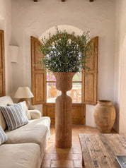A large woven rattan vase holding a tall green olive tree arrangement, styled in a rustic Mediterranean living room with a cream sofa, wooden shutters, and a terracotta pot.