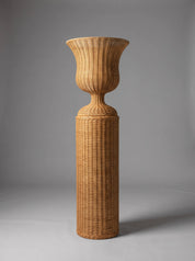 A tall, slender woven rattan stand or vase with a flared top and cylindrical base, photographed against a smooth gray studio background.