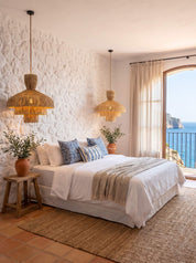A beautifully styled bedroom featuring a bed with white linens and blue patterned pillows, set against a textured white stucco wall with two woven pendant lights. A large arched window reveals a scenic view of the blue sea, and the room is decorated with potted plants and a natural jute rug.