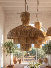 A large tiered pendant light made of natural woven straw or raffia hangs in a bright, sunlit room with white walls, a window, and potted plants. A second matching light is visible behind it.