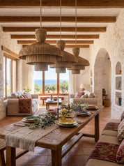 A rustic dining room with a long wooden table set with plates and greenery, three large woven pendant lights hanging from exposed wooden ceiling beams, and stone walls with built-in shelves in the background.