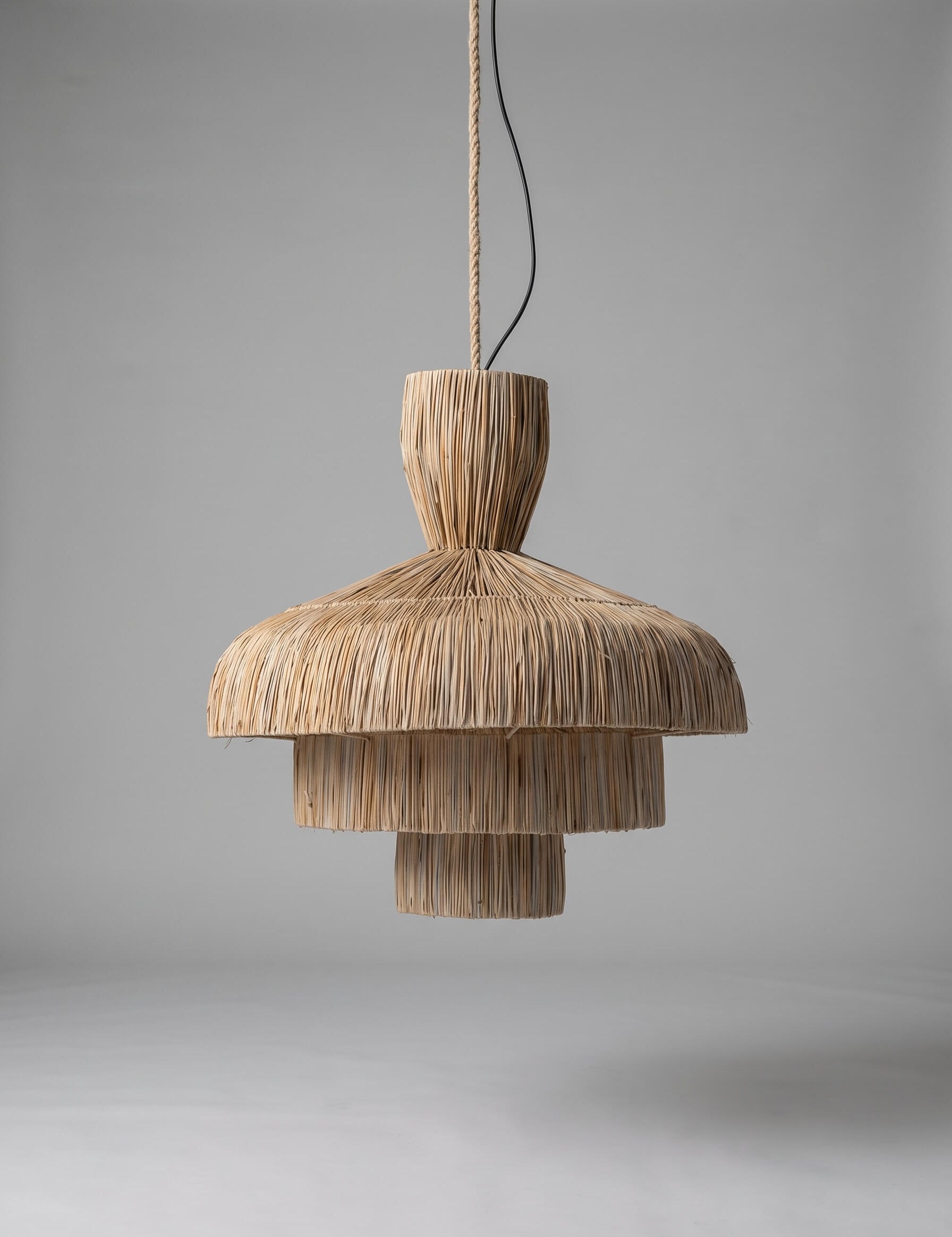 A natural woven reed pendant light with a tiered, layered design hanging from a thin cord against a neutral gray studio background.