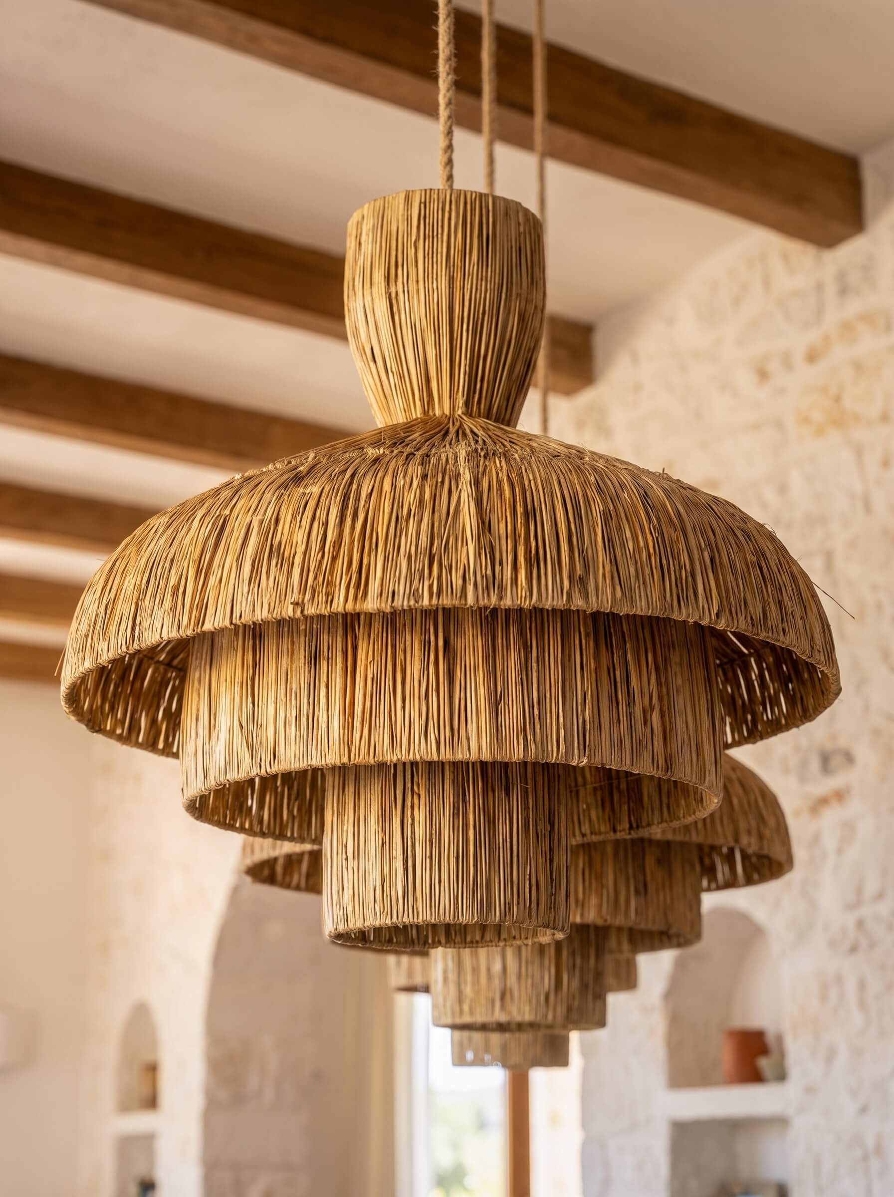 A large, tiered pendant light made from natural woven straw or raffia, hanging in a rustic room with stone walls and wooden ceiling beams.
