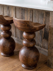 A close-up of a rustic wooden bar stool with a carved bowl-shaped seat and stacked round legs, shown in a warm indoor setting with wood flooring and a wooden wall panel background.