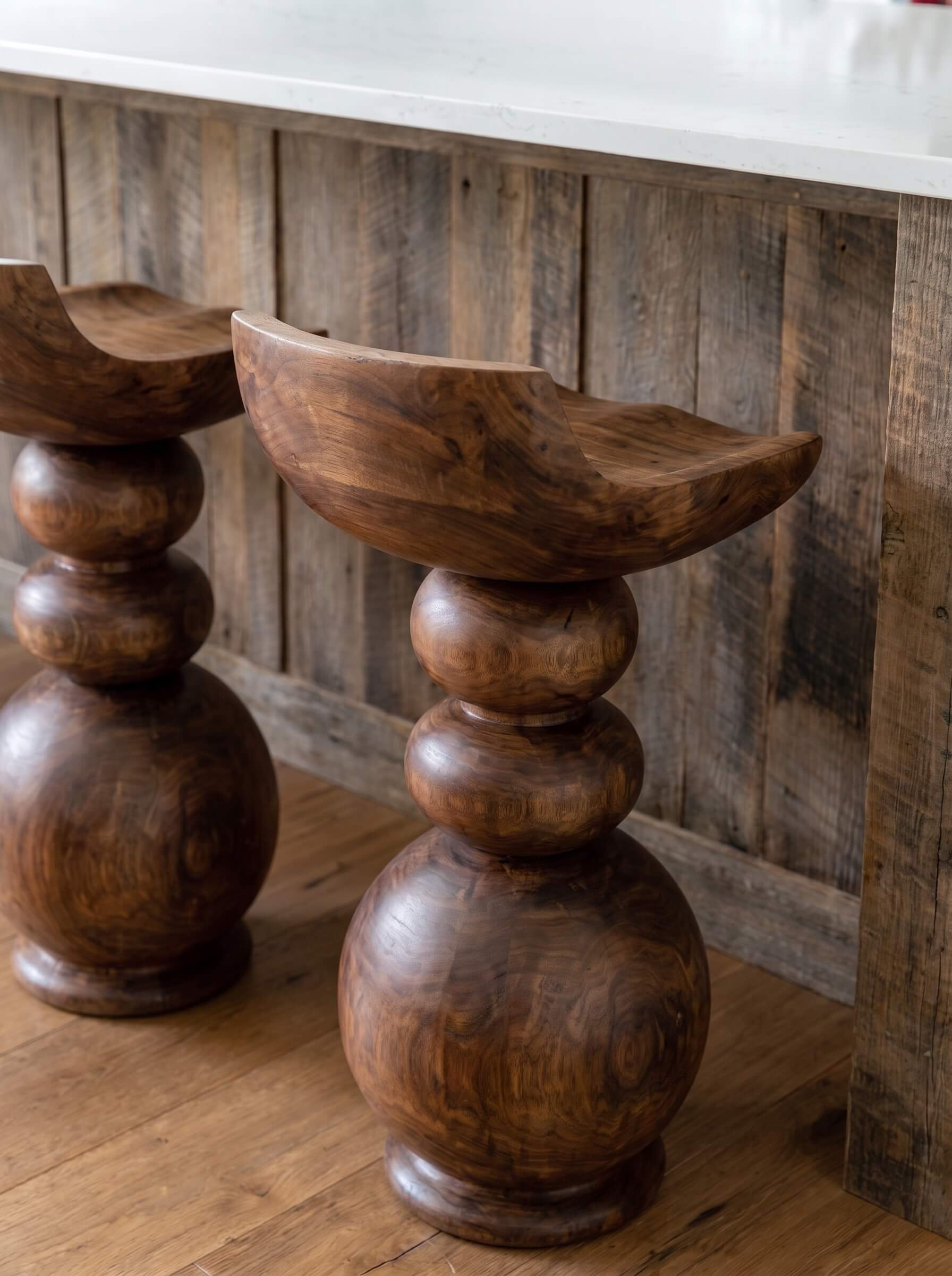 A close-up of a rustic wooden bar stool with a carved bowl-shaped seat and stacked round legs, shown in a warm indoor setting with wood flooring and a wooden wall panel background.