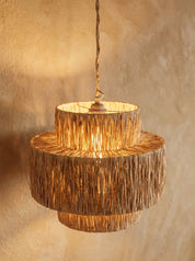 A tiered pendant light fixture made from natural woven reed or straw material, glowing with warm light against a textured beige plaster wall.
