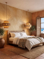 A rustic wooden bed dressed in white linens and a patterned throw blanket, styled in a warm, earthy bedroom with textured beige walls, two woven pendant lights, terracotta tiles, and a window with shutters.