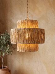 A tiered pendant light with a textured, woven natural fiber shade, glowing with warm light against a textured beige plaster wall with a potted plant in the background.