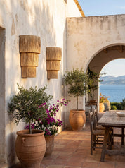 A rustic outdoor patio with terracotta pots, potted olive trees, a wooden dining table and chairs, and a white stucco wall with woven wall sconces. An arched doorway in the background reveals the blue sea and distant mountains under a clear sky.