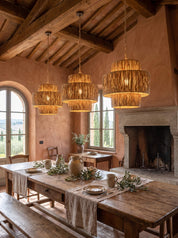 Three woven natural-fiber pendant lights hang over a rustic wooden dining table in a warm, earth-toned room with exposed wood beams, a stone fireplace, and large windows.