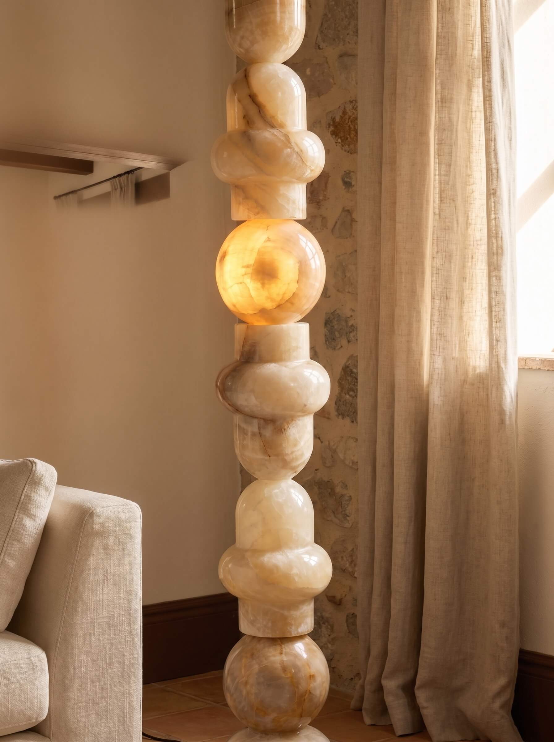 A tall sculptural floor lamp made of stacked polished stone spheres in warm cream and beige tones, lit from within near the center, standing in a softly styled room with curtains, a sofa edge, and patterned wallpaper.