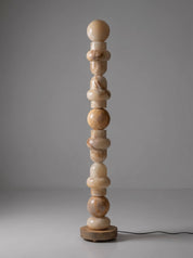 A tall, sculptural floor lamp made of stacked organic-shaped stone beads in natural beige and light brown tones, standing on a round wooden base against a neutral gray studio background.