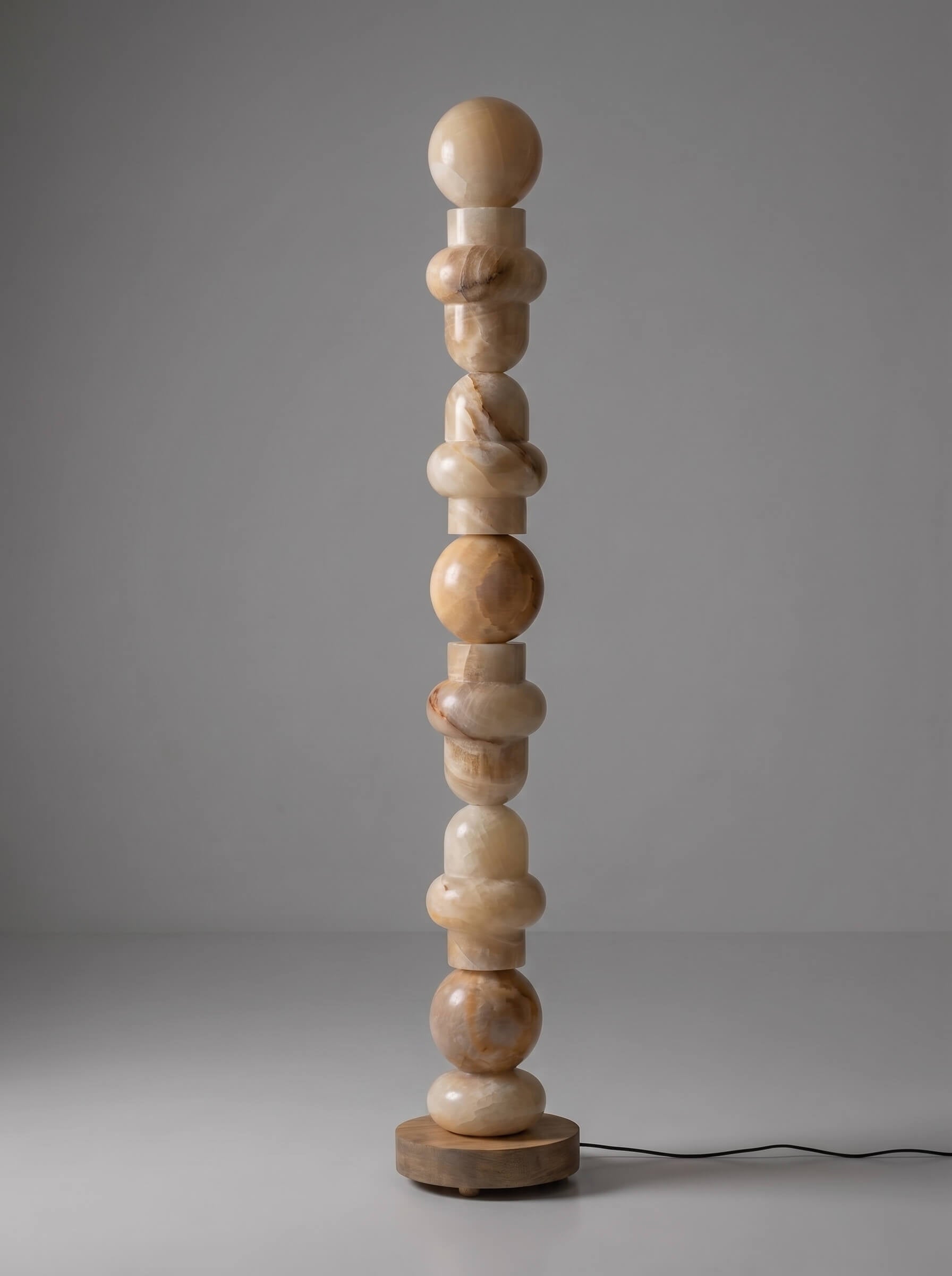 A tall, sculptural floor lamp made of stacked organic-shaped stone beads in natural beige and light brown tones, standing on a round wooden base against a neutral gray studio background.