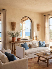 A bright, sunlit living room with a cream slipcovered sofa, matching armchair, rustic wood coffee table, blue accent pillows, terracotta planters, and an arched window overlooking the ocean.