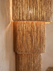 A close-up of a wall-mounted light fixture with three stacked cylindrical shades made of vertically textured natural fiber, glowing warm yellow against a beige plaster wall.