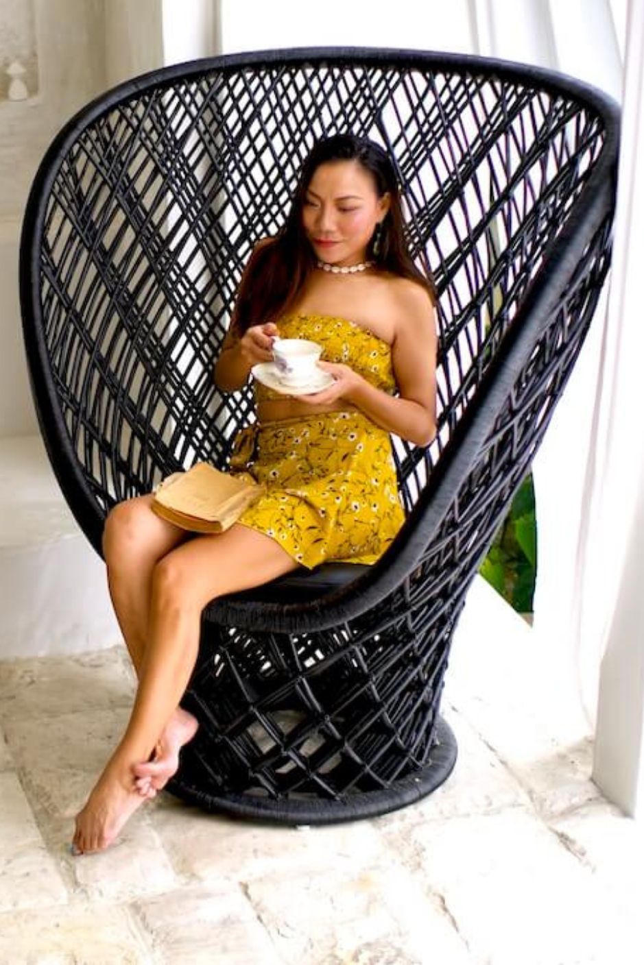 Woman in a yellow dress sitting in a black wicker chair holding a cup and saucer.