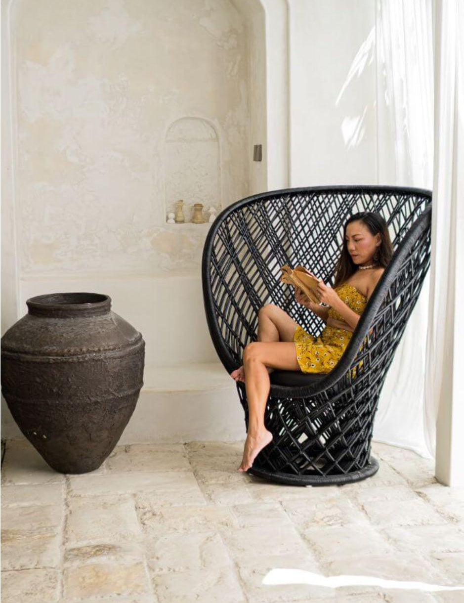 Woman sitting in a black wicker chair in a room with a large vase and white walls.