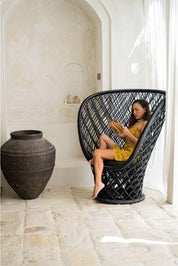 Woman sitting in a black wicker chair in a room with a large vase and white walls.
