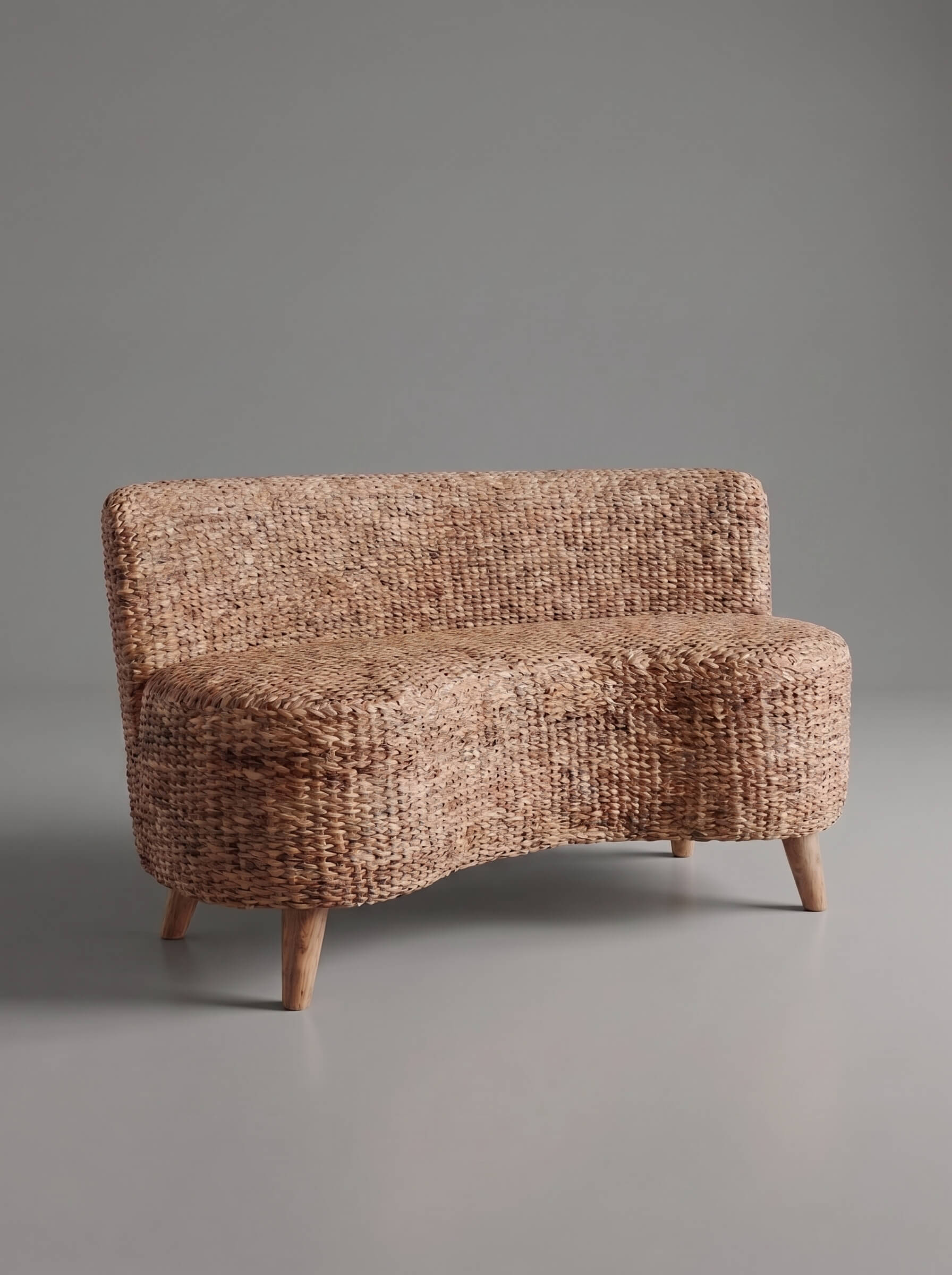 A modern curved upholstered bench or sofa with a tightly woven natural fiber texture in warm tan and brown tones, supported by four tapered wooden legs, set against a solid gray studio background.