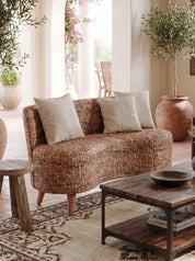 A curved woven rattan loveseat sofa with three neutral beige throw pillows, styled in a warm, sunlit room with a wooden coffee table, patterned rug, and potted olive trees.