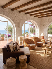 A bright, sunlit living room with white stone walls, exposed wooden ceiling beams, and large arched windows overlooking the sea. The space is furnished with a dark brown sofa, two woven wicker armchairs with cream cushions, a wooden coffee table, lamps, and natural fiber rugs.