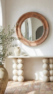 Round wooden mirror on a white wall above a stone console table with decorative stones.