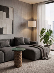A modern living room scene featuring a dark gray sectional sofa with matching throw pillows, a textured green marble-top side table, a beige floor lamp, and a large potted monstera plant in front of a window with sheer curtains.
