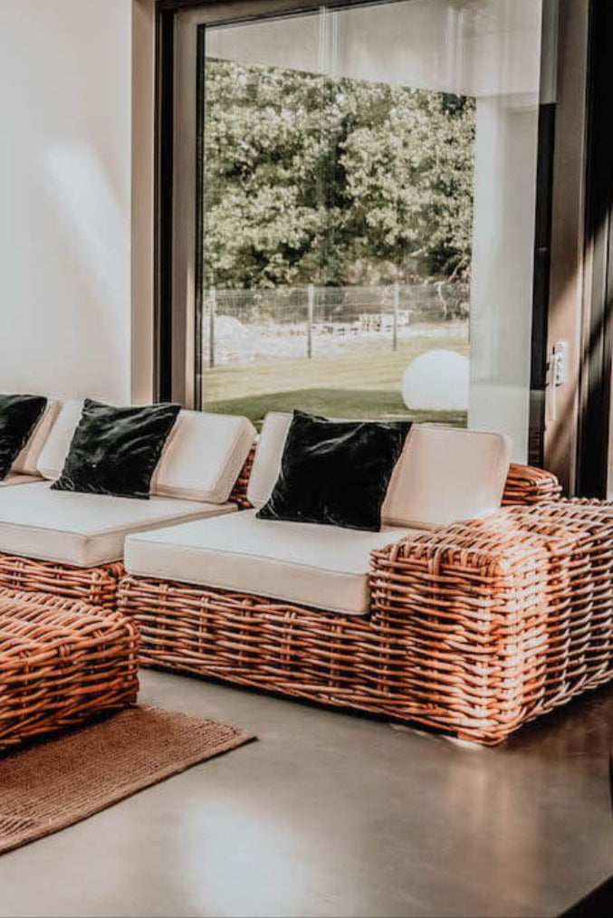 Wicker outdoor sofa with beige cushions and black pillows in a room with large windows.