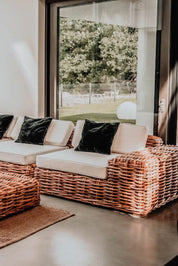 Wicker outdoor sofa with beige cushions and black pillows in a room with large windows.
