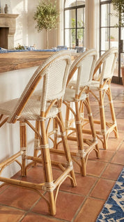 Rattan bar stools in front of a wooden counter with a tiled floor and large windows in the background.