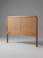 A modern wooden bed headboard with a natural cane or rattan woven center panel and a light oak wood frame, shown against a neutral gray studio background.