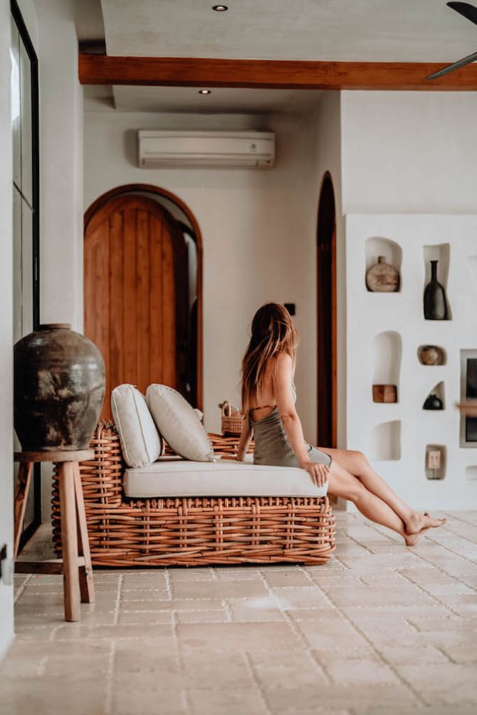Woman sitting on a wicker chair in a room with a wooden door and decorative wall shelves.