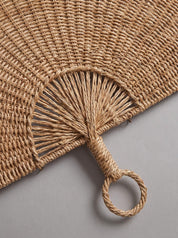 Close-up of a natural woven straw fan with a loop handle, showing the detailed texture of the material on a neutral gray background.