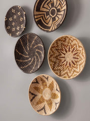 Five handwoven basket wall decor pieces with various geometric and spiral patterns in natural tan, brown, and black, arranged on a neutral gray background.