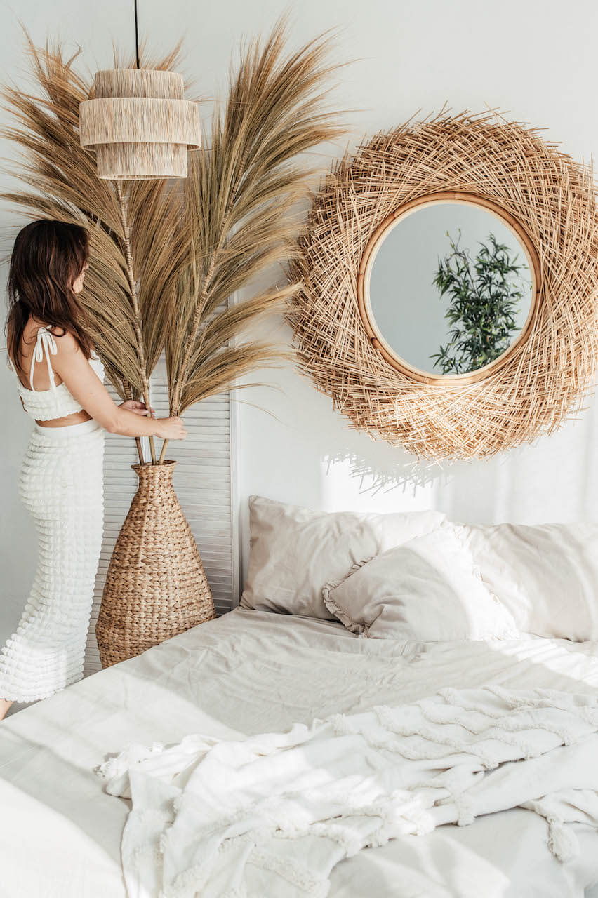 Woman arranging pampas grass in a woven vase next to a bed with a round mirror above.