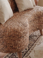 Close-up of a woven natural-fiber upholstered bench or sofa corner with cream throw pillows and a patterned rug in a warm indoor setting.