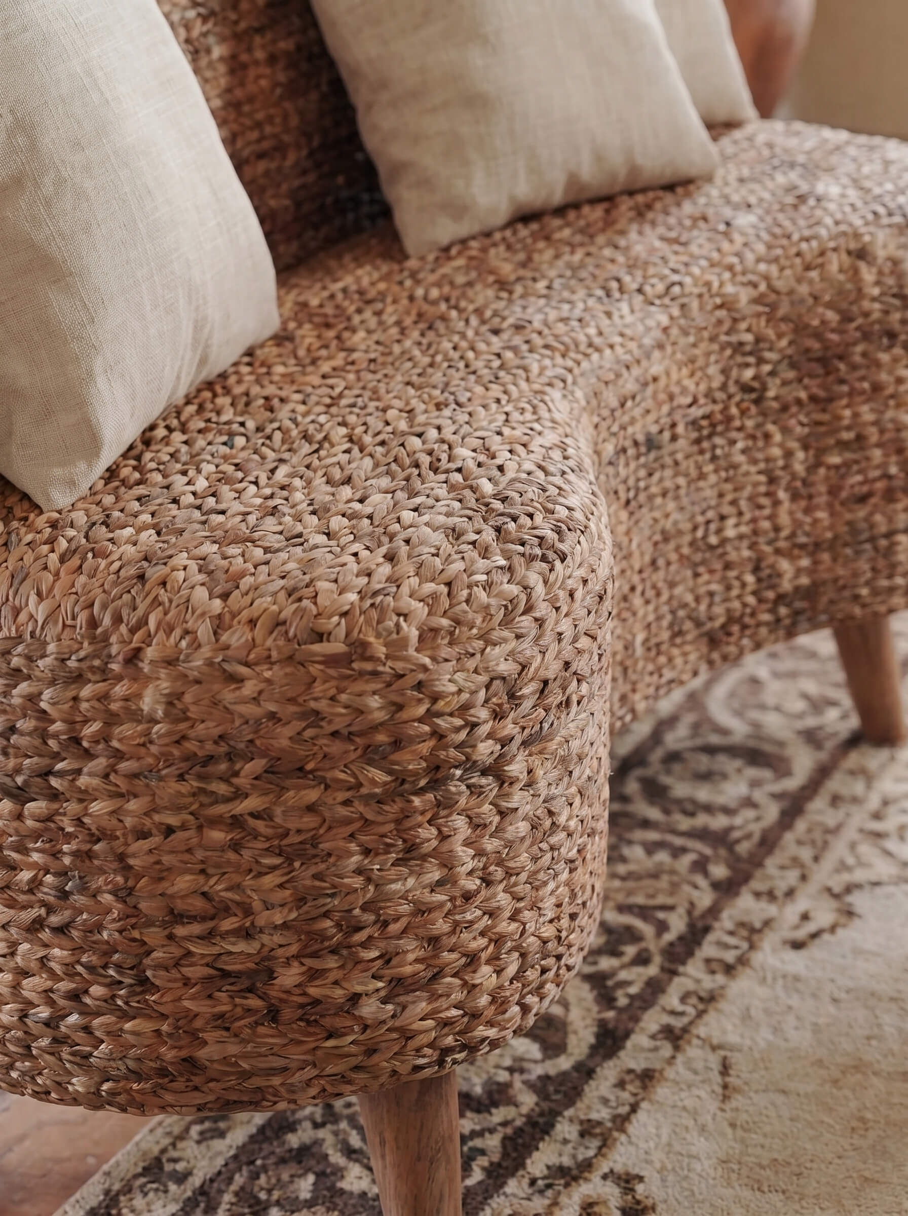 Close-up of a woven natural-fiber upholstered bench or sofa corner with cream throw pillows and a patterned rug in a warm indoor setting.
