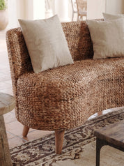 A curved woven seagrass sofa with natural wood legs, styled with two beige throw pillows in a bright, sunlit room.
