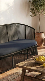 Black metal bench with dark cushion in a room with a wooden table and plant.