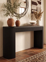 A modern black console table with a fluted front and open lower compartment, styled in a warm interior with terracotta vases, a round mirror, books, and dried grasses on a tiled floor.