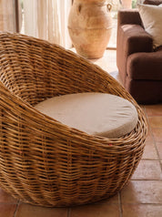 A natural woven rattan armchair with a round beige cushion, set in a warm, rustic room with terracotta tile flooring and a brown sofa in the background.