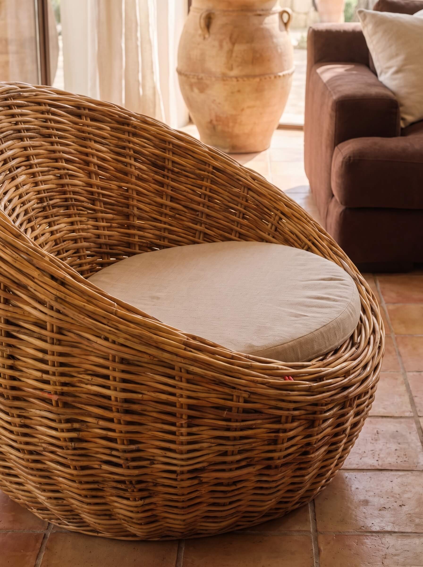 A natural woven rattan armchair with a round beige cushion, set in a warm, rustic room with terracotta tile flooring and a brown sofa in the background.