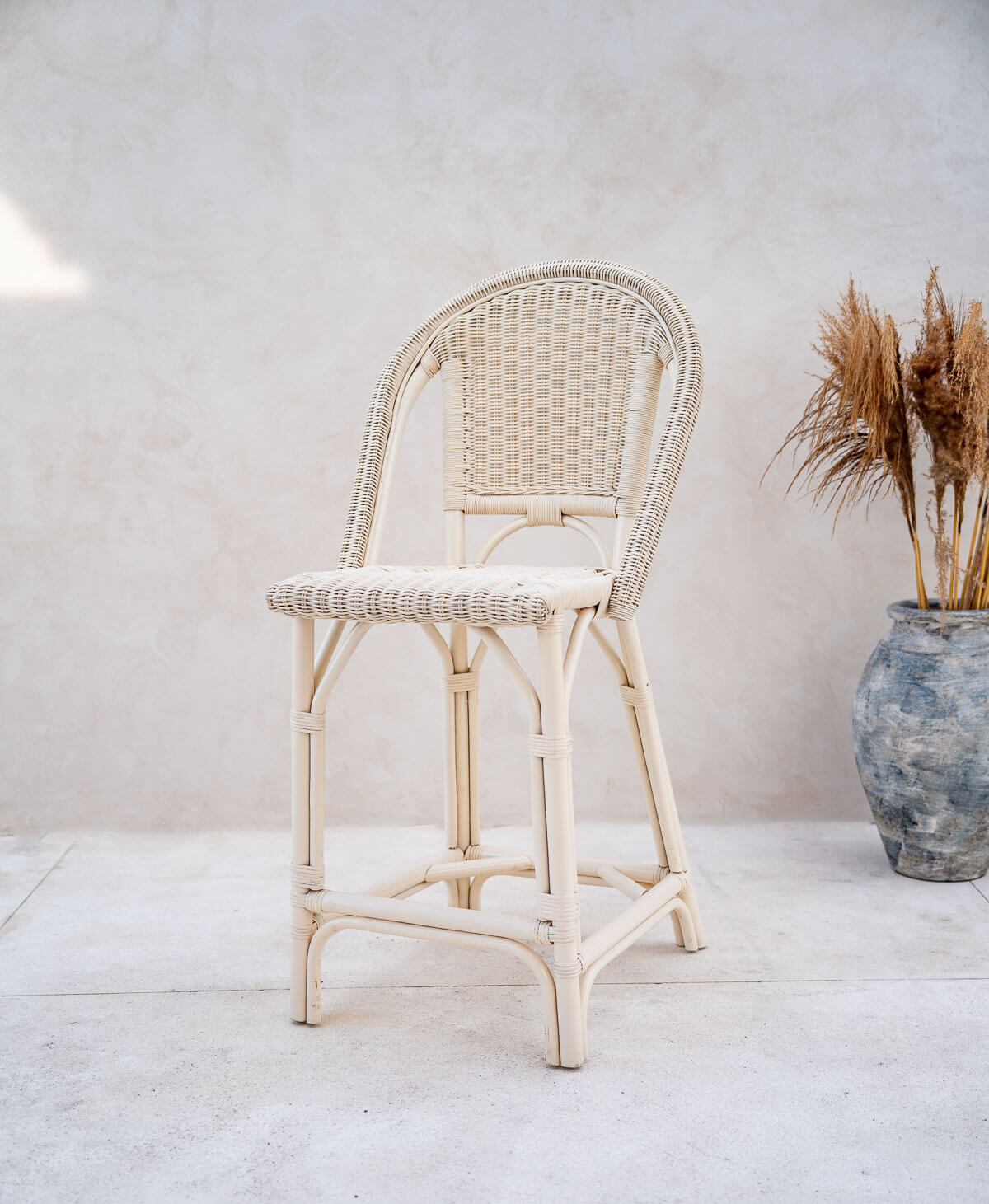 Wicker bar stool against a white wall with a vase of dried plants on the right.