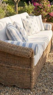 Wicker outdoor sofa with white cushions and blue striped pillows in a garden setting.