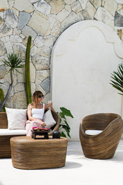 Woman sitting on a wicker chair in an outdoor setting with stone wall and plants.