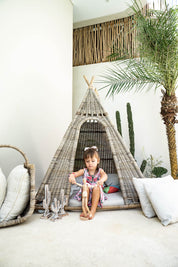 Child sitting inside a woven teepee in a room with a palm tree and decorative plants.