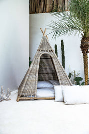 Wicker teepee with white bedding against a white wall with decorative plants.