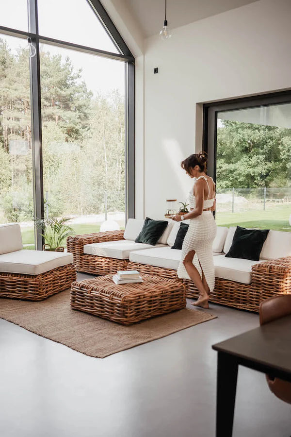 Woman standing in a modern living room with wicker furniture and large windows.