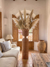 A tall woven basket vase filled with dried pampas grass in a rustic living room setting with a sofa, wooden shutters, and a wooden table.