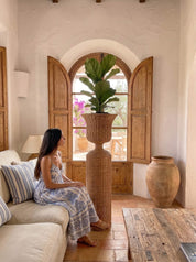 A woman sits on a cream sofa in a rustic, sunlit room with white plaster walls, wooden shutters, and a tall woven planter holding a green plant in the center.